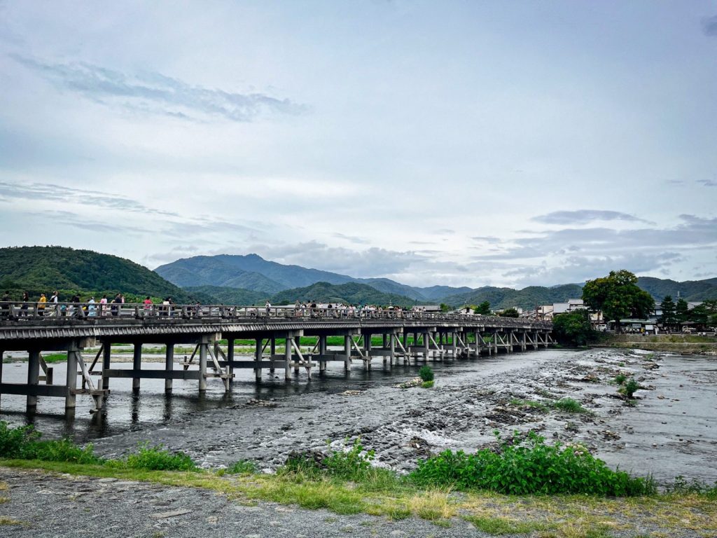 嵐山の渡月橋を岸から撮影。橋には大勢の人が渡っている。空は曇り空。