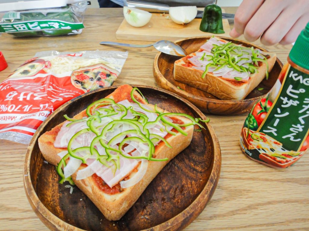 トーストにピザソースやベーコン、ピーマンなどを載せている様子。